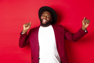 Noel ve insanlar kavramı. Mutlu siyah adam yeni yılı kutluyor ve dans ediyor, parti kıyafeti giyiyor, kırmızı arka plan