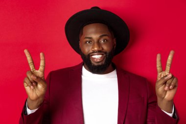 Yakışıklı, sakallı, barış işaretleri gösteren, mutlu gülümseyen, kırmızı arka planda duran siyah adamın yakın çekimi.