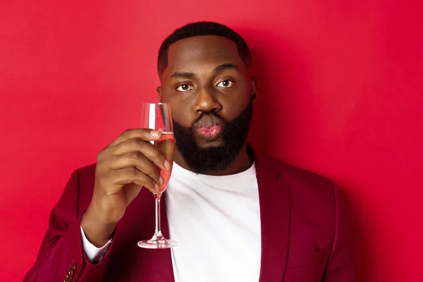 Close-up of funny Black man tasting champagne from glass, looking silly and pucker lips, celebrating new year, standing against red background