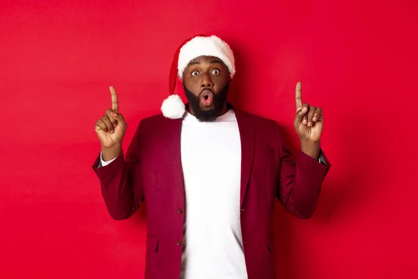 Concepto de Navidad, fiesta y vacaciones. Impresionado hombre negro mostrando oferta, señalando los dedos hacia arriba en el logotipo, diciendo wow, de pie sobre el fondo rojo — Foto de Stock