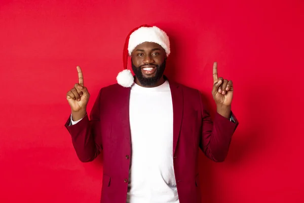 Concepto de Navidad, fiesta y vacaciones. Hombre afroamericano guapo en sombrero de santa sonriendo, señalando con los dedos hacia arriba y mostrando publicidad, de pie sobre el fondo rojo — Foto de Stock
