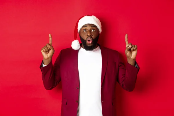 Concepto de Navidad, fiesta y vacaciones. Impresionado hombre negro en sombrero de santa señalando los dedos hacia arriba, diciendo wow y mirando fijamente a la cámara, echa un vistazo a la oferta promocional, fondo rojo — Foto de Stock