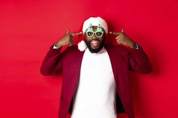 Merry Christmas. Cheerful Black man wearing funny party glasses and santa hat, smiling joyful, celebrating winter holidays, standing over red background