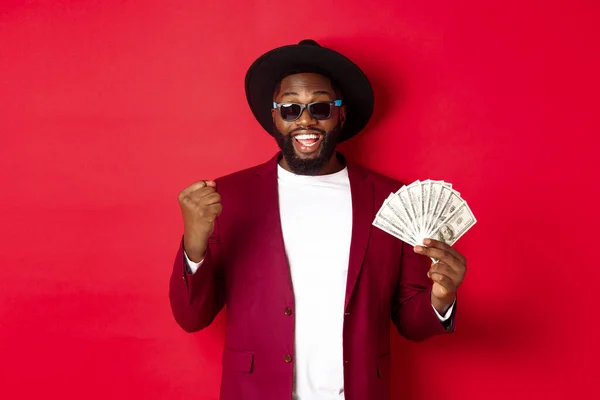 Cheerful african american guy in sunglasses, black hat and blazer, winning prize money, holding dollars and looking satisfied, standing over red background
