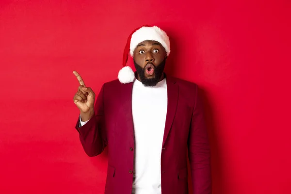 Concepto de Navidad, fiesta y vacaciones. Impresionado hombre negro con barba, vestido con sombrero de santa, señalando con el dedo hacia la izquierda y jadeando sorprendido, de pie sobre fondo rojo — Foto de Stock