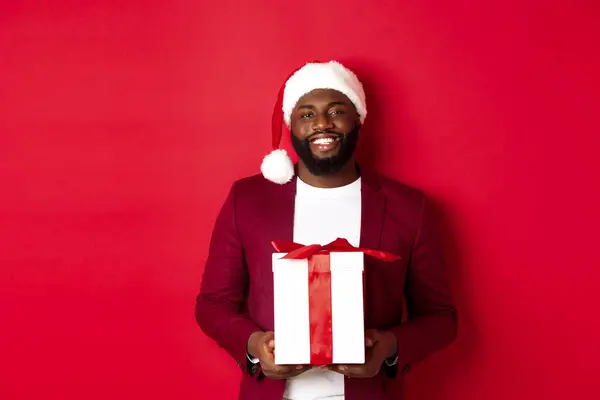 Navidad, Año Nuevo y concepto de compras. Feliz barbudo afroamericano hombre con la celebración de Navidad presente, sonriendo a la cámara, de pie en sombrero de santa sobre fondo rojo — Foto de Stock