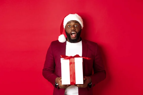 Navidad, Año Nuevo y concepto de compras. Emocionado hombre negro recibiendo regalo de Navidad, mirando sorprendido, sosteniendo regalo envuelto y mirando fijamente a la cámara, usando sombrero de santa — Foto de Stock