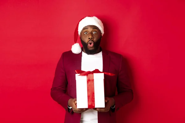 Navidad, Año Nuevo y concepto de compras. Sorprendido afroamericano hombre en sombrero de santa diciendo wow, celebración de regalo de vacaciones y mirando a la cámara sorprendido, fondo rojo — Foto de Stock
