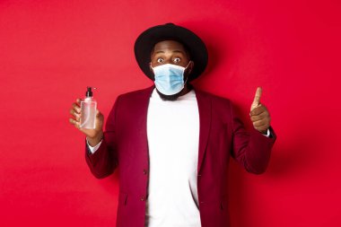 Covid-19, karantina ve tatil konsepti. Etkilenmiş siyah adam parti kıyafeti ve tıbbi maske takıyor, baş parmağını kaldırıyor ve el dezenfektanını gösteriyor, kırmızı arka planda duruyor.