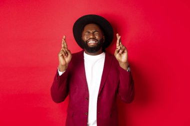 Umut dolu Afrikalı Amerikalı bir adam dilek tutuyor, şans diliyor ve iyimser gülümsüyor, kırmızı parti geçmişine karşı duruyor.