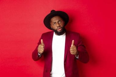 Etkileyici siyahi adam iyi iş çıkardığını söylüyor, başparmağınla mutlu ol, bir şeyleri öv, kırmızı arka planda dur diyor.