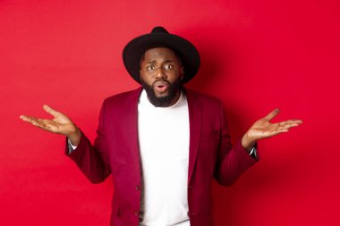 Kafası karışmış ve endişeli siyah adam ellerini yana açmış, kameraya şaşkın şaşkın bakıyor, neyin yanlış olduğunu soruyor, kırmızı arka planda duruyor.