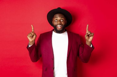 Kış tatili ve alışveriş konsepti. Neşeli siyahi adam yukarı bakıyor ve işaret ediyor, Xmas tanıtımına bakıyor, kırmızı arka planda duruyor.