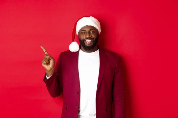 Concepto de Navidad, fiesta y vacaciones. Hombre negro guapo en sombrero de santa señalando el dedo a la izquierda, mostrando publicidad, de pie feliz sobre fondo rojo — Foto de Stock