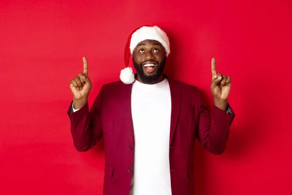 Concepto de Navidad, fiesta y vacaciones. Feliz hombre negro mirando la promoción de Año Nuevo, señalando con los dedos hacia arriba y sonriendo, de pie sobre el fondo rojo — Foto de Stock