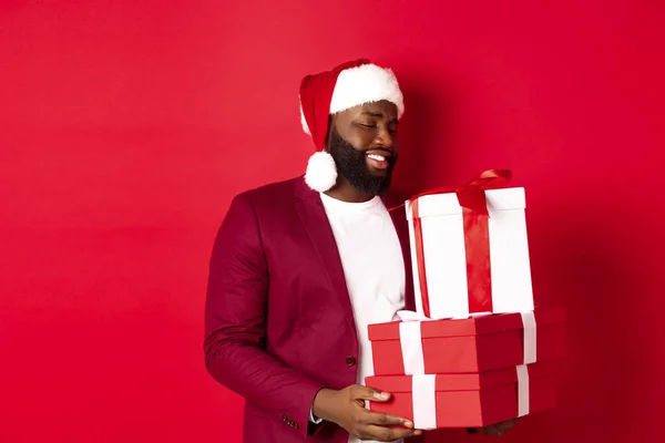 Navidad, Año Nuevo y concepto de compras. Feliz africano amaerican hombre buscando tocado y agradecido en regalos de Navidad, recibir regalos, usando sombrero de santa, fondo rojo — Foto de Stock