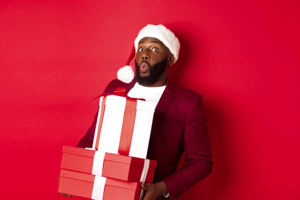 Navidad, Año Nuevo y concepto de compras. Feliz hombre negro en sombrero de santa y blazer celebración de regalos de Navidad, traer regalos y sonreír, de pie sobre fondo rojo — Foto de Stock