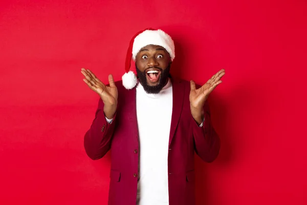 Concepto de Navidad, fiesta y vacaciones. Sorprendido y feliz hombre afroamericano en sombrero de santa, mirando con regocijo a la cámara, de pie sobre fondo rojo — Foto de Stock