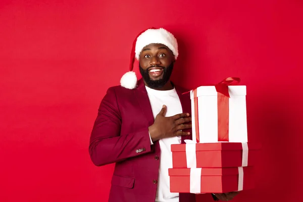 Navidad, Año Nuevo y concepto de compras. Feliz hombre negro recibiendo regalos de Navidad, diciendo gracias y sonriendo agradecido, de pie en sombrero de santa sobre fondo rojo — Foto de Stock