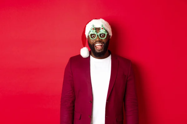 Merry Christmas. Cheerful Black man wearing funny party glasses and santa hat, smiling joyful, celebrating winter holidays, standing over red background