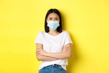 Covid, sağlık ve salgın hastalık konsepti. Beyaz tişörtlü ve tıbbi maskeli Asyalı kadın göğsünde çapraz kollar ve kamera karşısında üzgün ve hayal kırıklığına uğramış görünüyor.