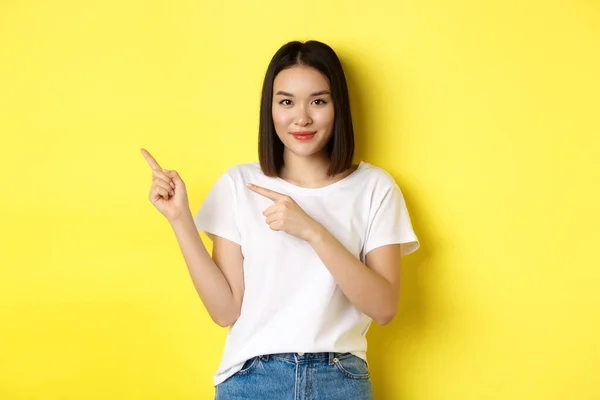 Concetto di bellezza e moda. Bella donna asiatica in t-shirt bianca che punta le dita a sinistra, in piedi su sfondo giallo — Foto Stock