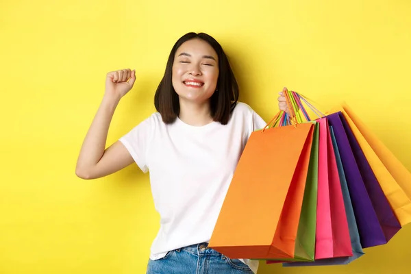 Щаслива азіатська жінка відчуває себе задоволеною після покупок у продажу, тримаючи паперові мішки та розтягуючись із задоволеною посмішкою, стоячи над жовтим тлом — стокове фото
