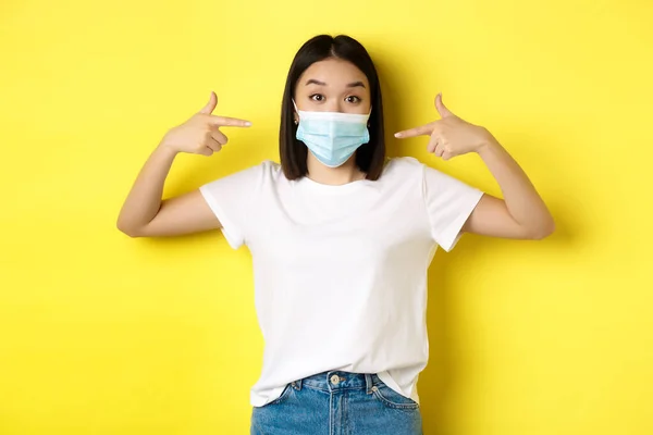 Ковид-19, концепция карантина и социального дистанцирования. Молодая азиатка в белой футболке, указывающая на свою медицинскую маску из коронавируса, желтый фон — стоковое фото