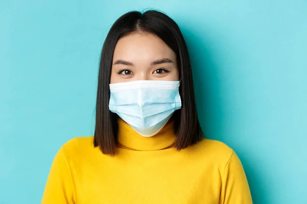 Covid-19, socialt avståndstagande och pandemikoncept. Närbild av ung asiatisk kvinna med kort mörkt hår, bär medicinsk mask och ler med ögon, ser hoppfull på kameran — Stockfoto