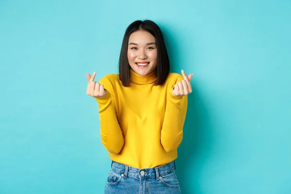 Menina asiática bonita em suéter amarelo, sorrindo e mostrando corações de dedo, de pé feliz contra o fundo azul — Fotografia de Stock