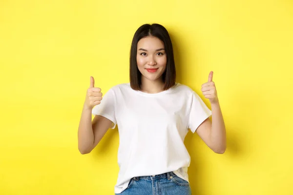 Όμορφη νεαρή Ασιάτισσα γυναίκα σε λευκό t-shirt, δείχνει τους αντίχειρες επάνω και χαμογελώντας, επαίνους καλή προσφορά, προτείνουμε προϊόν, στέκεται ικανοποιημένοι πάνω από κίτρινο φόντο — Φωτογραφία Αρχείου