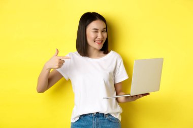 E-ticaret ve alışveriş konsepti. Güzel görünümlü Asyalı kız kameraya göz kırpıyor, gülümsüyor ve bir dizüstünü işaret ediyor, bir şey gösteriyor, sarı arka planda duruyor.