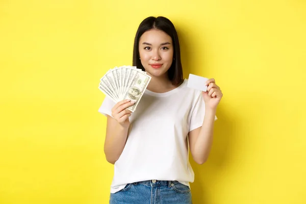 Vacker asiatisk kvinna med kort mörkt hår, bär vit t-shirt, visar pengar i dollar och plast kreditkort, står över gul bakgrund — Stockfoto