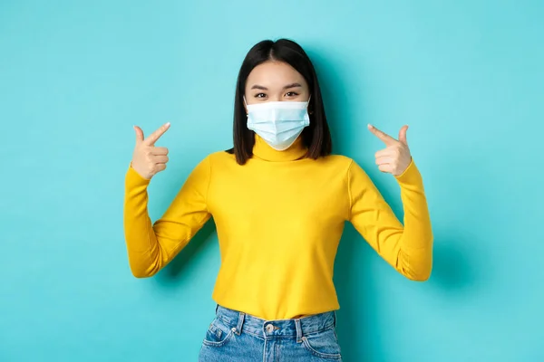 Covid-19, socialt avståndstagande och pandemikoncept. Ung asiatisk kvinna skydda sig från coronavirus, peka finger åt hennes medicinska mask, står över blå bakgrund — Stockfoto