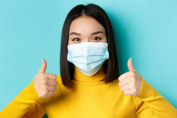 Covid-19, socialt avståndstagande och pandemikoncept. Närbild av ung asiatisk kvinna i medicinsk mask visar tummen upp, ge godkännande, beröm bra erbjudande, står över blå bakgrund — Stockfoto