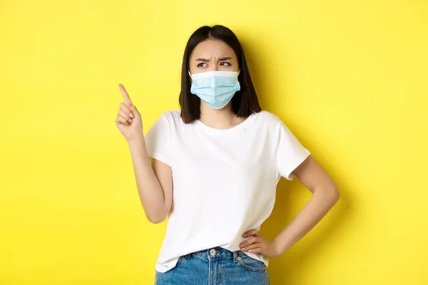 Ковид, здравоохранение и пандемия. Азиатка в медицинской маске и белой футболке указывая пальцем на логотип верхнего левого угла, показывая продвижение, желтый фон — стоковое фото