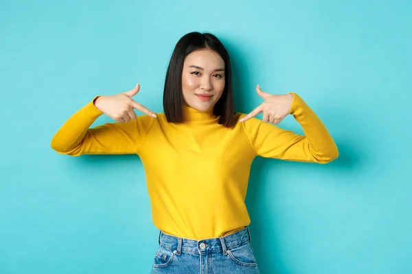 Självsäker asiatisk kvinna i snygg tröja pekande fingrar på logotypen på mitten, ler bestämd mot kameran, står över blå bakgrund — Stockfoto