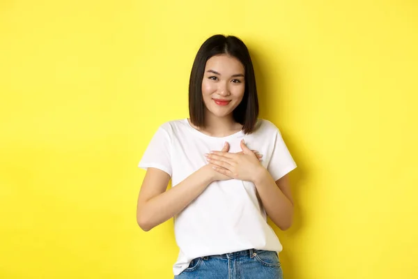 Güzellik ve moda konsepti. Yürekten Asyalı bir kız, kalbi ile el ele tutuşuyor ve gülümsüyor dokunuyor, teşekkür ediyor, sarı arka planda duruyor. — Stok fotoğraf