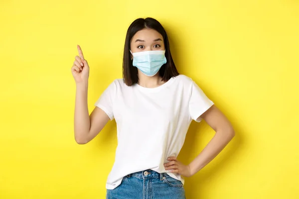 Covid, sjukvård och pandemi koncept. Asiatisk kvinnlig modell i medicinsk mask och vit t-shirt pekar finger på övre vänstra hörnet logotyp, visar befordran, gul bakgrund — Stockfoto