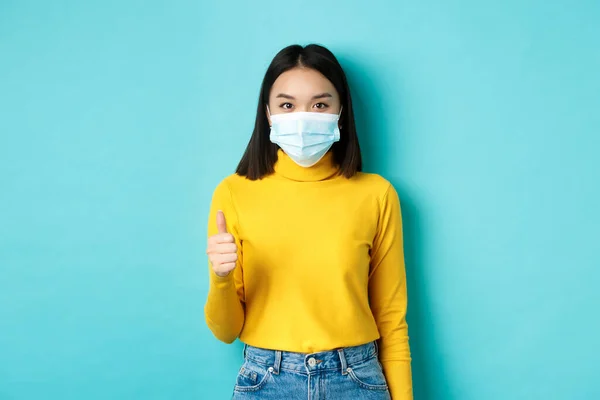 Covid-19, κοινωνική αποστασιοποίηση και πανδημία. Νεαρή γυναίκα με ιατρική μάσκα δείχνει τους αντίχειρες της σε επιδοκιμασία, λέγοντας ναι, στέκεται πάνω από το μπλε φόντο — Φωτογραφία Αρχείου