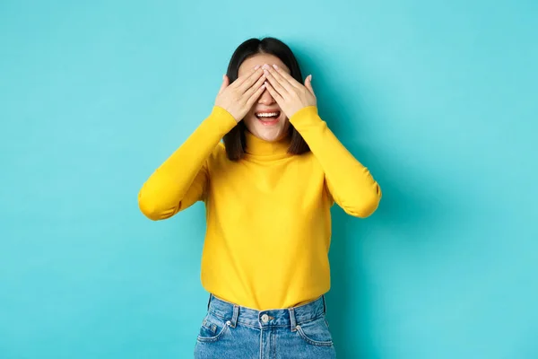 Cheerful asian girl in yellow pullover waiting for surprise, playing hide n seek and smiling, expecting gift with eyes closed, standing over blue background — Stock Photo, Image