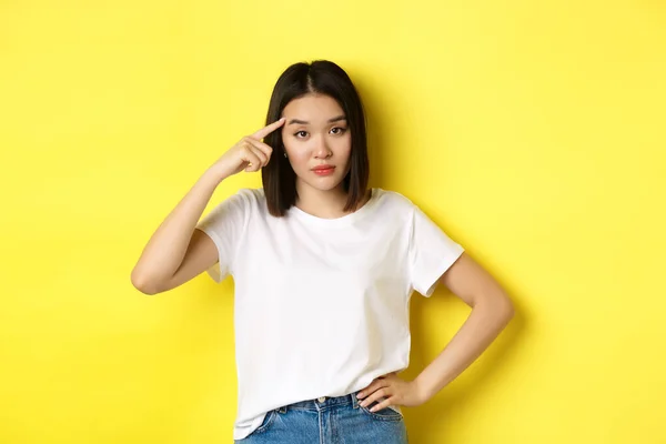 Sinirli genç Asyalı bir kadın aptal birini azarlıyor, parmağıyla kafasını işaret ediyor, deli misin diye soruyor, sarı arka planda dikiliyor. — Stok fotoğraf