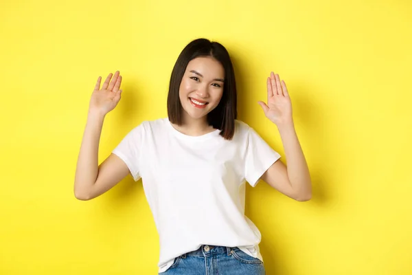 친절 한 젊은 아시아 여자가 인사를 하고, 빈 손을 들고 미소를 짓고, 노란 배경 위에 서서 인사를 합니다. — 스톡 사진
