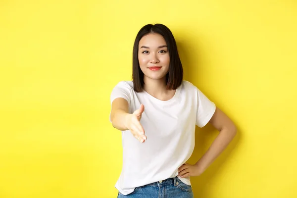 Αυστηρή Ασιάτισσα με λευκό μπλουζάκι, άπλωσε το χέρι της για χειραψία και χειρονομία χαιρετισμού, λέγοντας γεια, συστήνεται, στέκεται πάνω από κίτρινο φόντο — Φωτογραφία Αρχείου