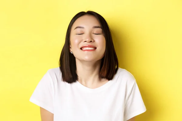 Lähikuva onnellinen ja rento aasialainen nainen nauttii auringosta, hymyilee silmät kiinni ja näyttää iloinen, seisoo keltainen tausta — kuvapankkivalokuva
