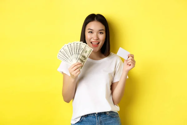 Красивая азиатская женщина с короткими темными волосами, в белой футболке, с деньгами в долларах и пластиковой кредиткой, стоящая на желтом фоне — стоковое фото