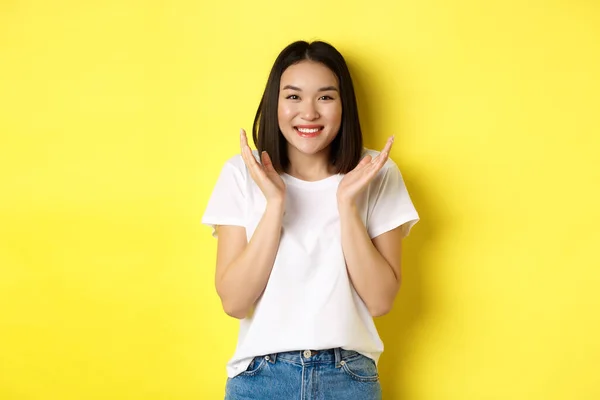 Ομορφιά και μόδα έννοια. Ενθουσιασμένη Ασιάτισσα χειροκροτεί και χαμογελά χαρούμενη στην κάμερα, στέκεται με λευκό μπλουζάκι σε κίτρινο φόντο — Φωτογραφία Αρχείου