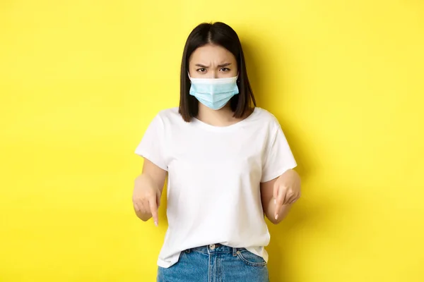 Covid-19，流行病和社会疏远概念。戴着医疗面具的令人失望的亚洲女孩，皱着眉头，用手指指着标识，站在黄色的背景上 — 图库照片