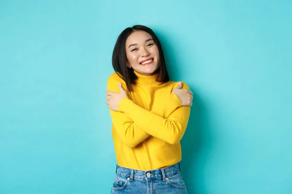 Menina bonita alegre abraçando o próprio corpo, sorrindo e abraçando-se, de pé despreocupado em pulôver amarelo contra fundo azul — Fotografia de Stock