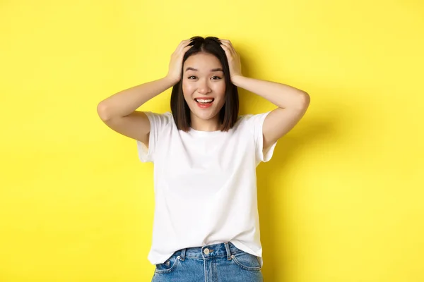Sorprendido chica asiática ganar premio y mirando con emoción e incredulidad, cogido de la mano en la cabeza y sonriendo feliz, de pie sobre el fondo amarillo — Foto de Stock